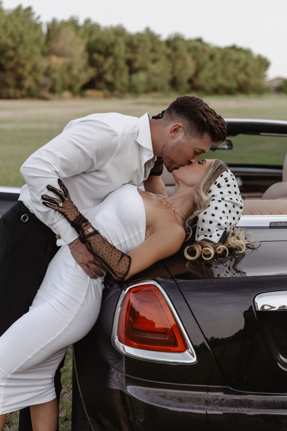 A couple shares a romantic kiss leaning against a black convertible car outdoors.