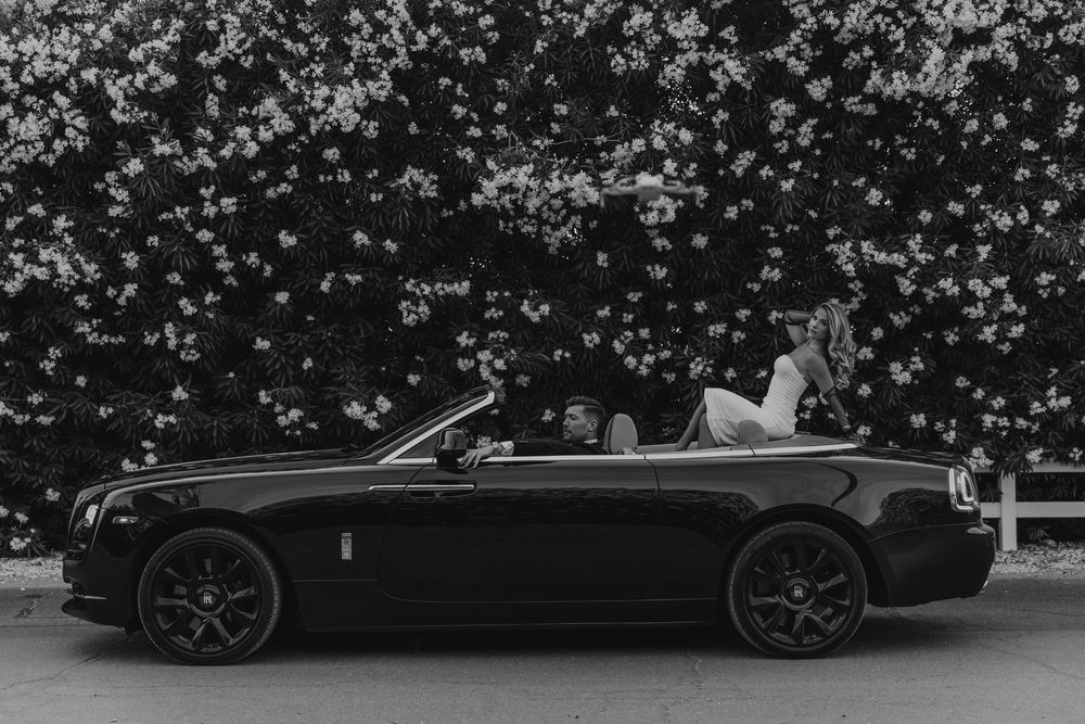 Black and white photo of a couple posing in a luxury Rolls-Royce convertible in front of a flowering hedge.