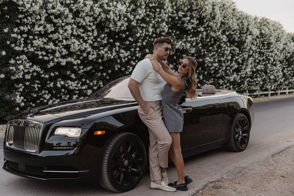 Couple poses romantically in front of a black Rolls-Royce luxury car near a white flowering hedge.
