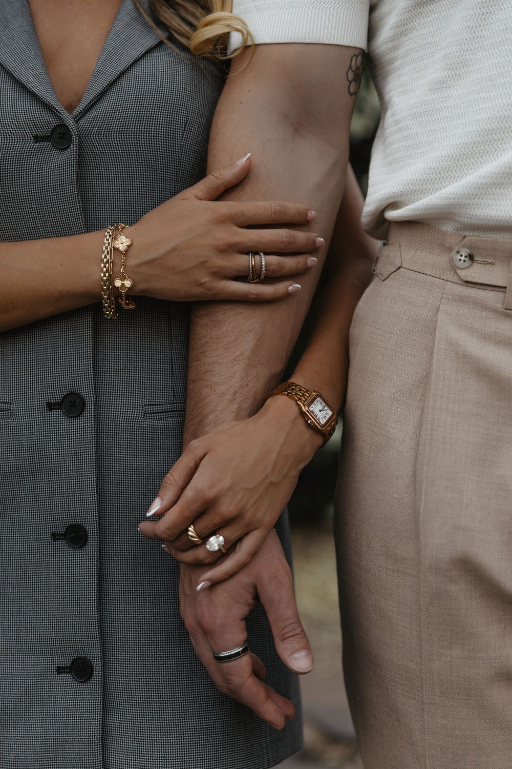 Close-up of couple holding hands showing gold bracelets, rings, and watch accessories outdoors.