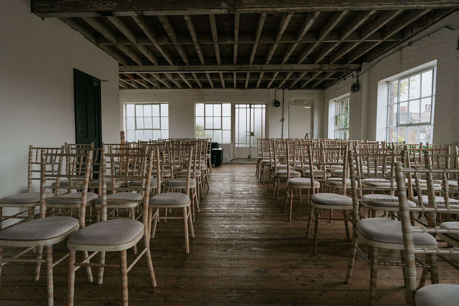 Empty wedding ceremony venue with rows of wooden chiavari chairs on hardwood floors and large windows in rustic space.