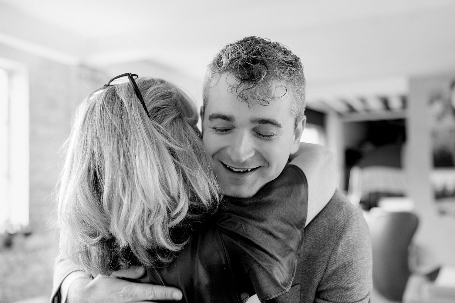 A young man smiles warmly while sharing a heartfelt hug with a woman in a bright indoor setting.