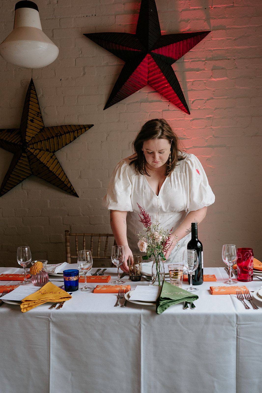 Woman in white dress arranging floral centerpiece on elegantly set dining table with colorful star decorations on brick wall.