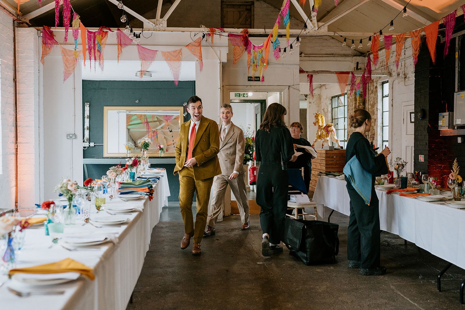 Colorful bunting decorates a bright event venue with long tables and guests mingling at a festive gathering.