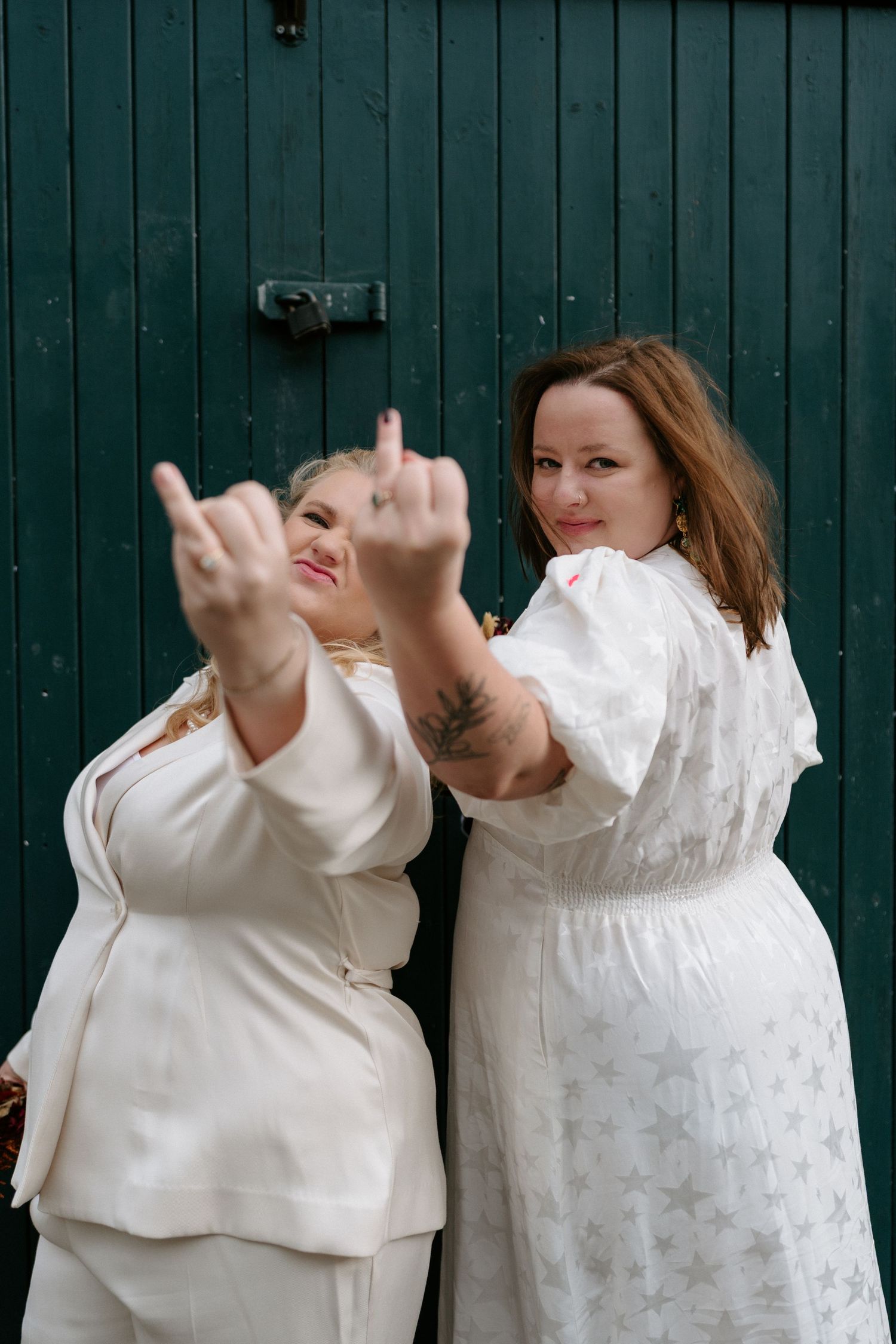 Two women in white wedding outfits playfully gesture at camera in front of a green wooden door.
