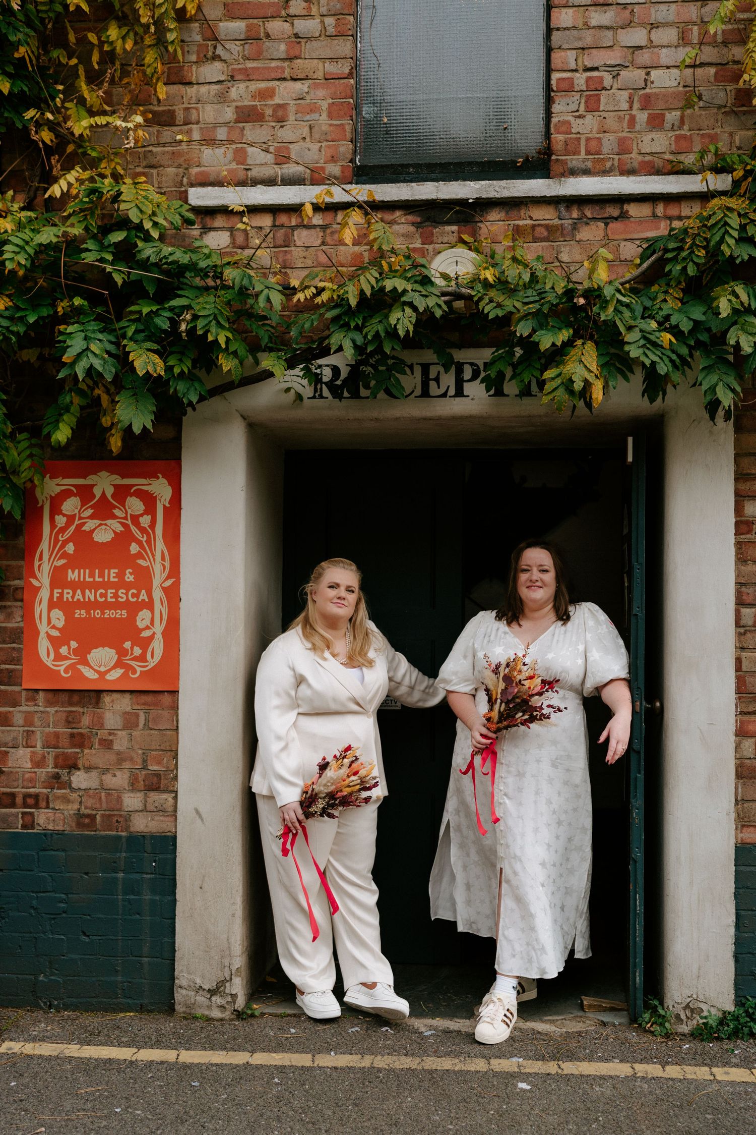 Two brides in white outfits holding bouquets standing at reception entrance of brick building.