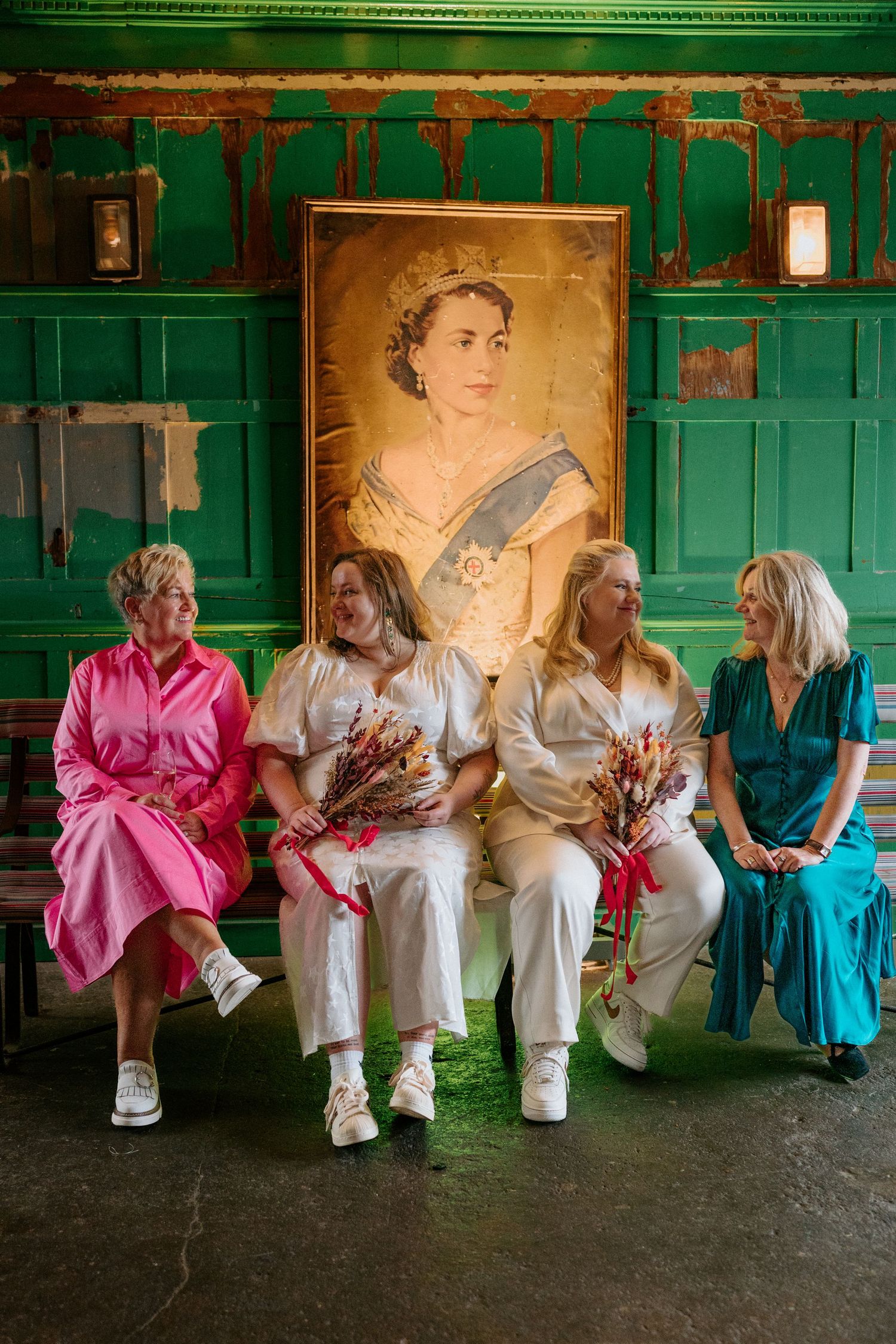 Four women in colorful dresses sit together holding bouquets beneath a regal portrait in a green room.