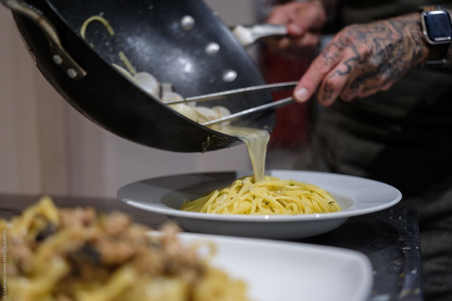 Chef with tattooed hands pouring creamy sauce over fresh spaghetti pasta in a white bowl.
