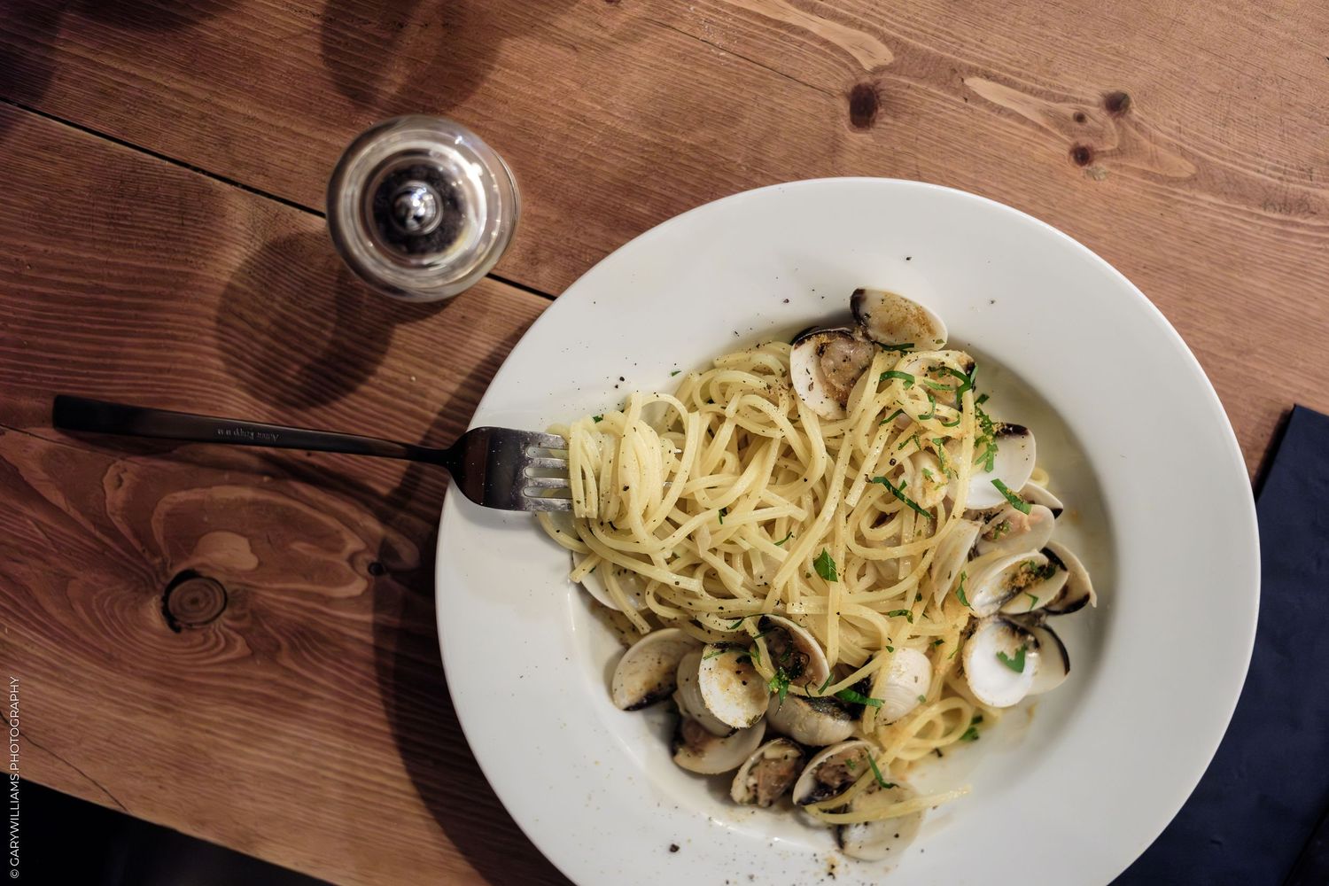 Spaghetti alle vongole with clams on white plate, fork, and pepper grinder on rustic wooden table.