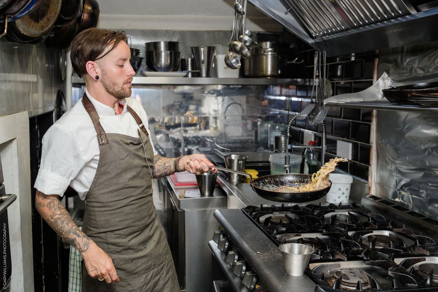 Tattooed chef in apron cooking in a professional kitchen, sauteing food in a pan on a gas stove.