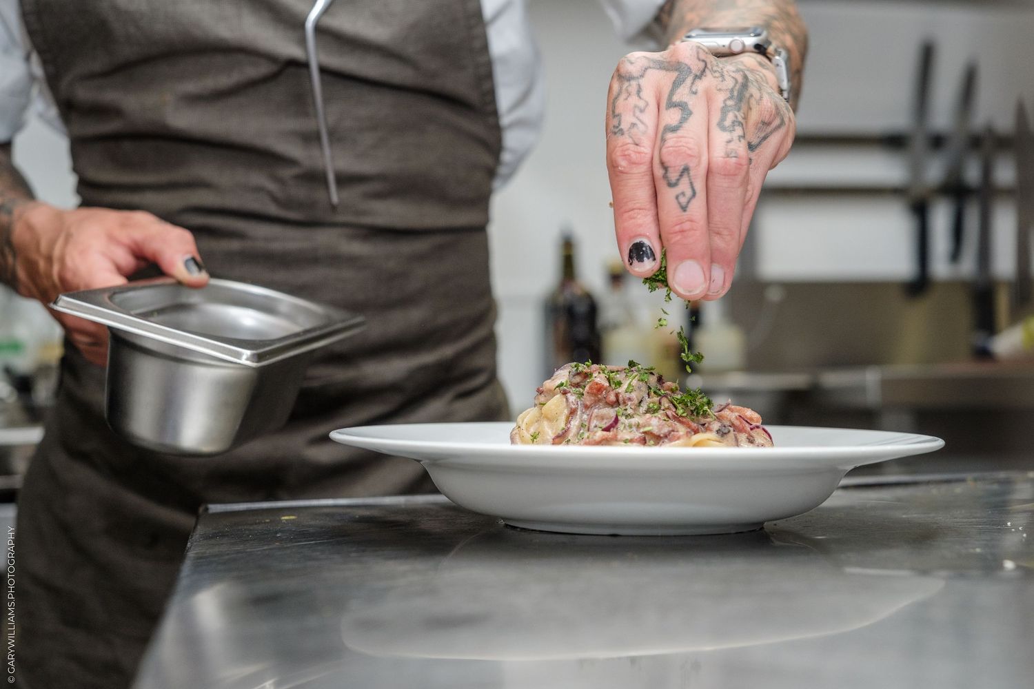 Tattooed chef garnishing a pasta dish on a white plate in a professional kitchen.