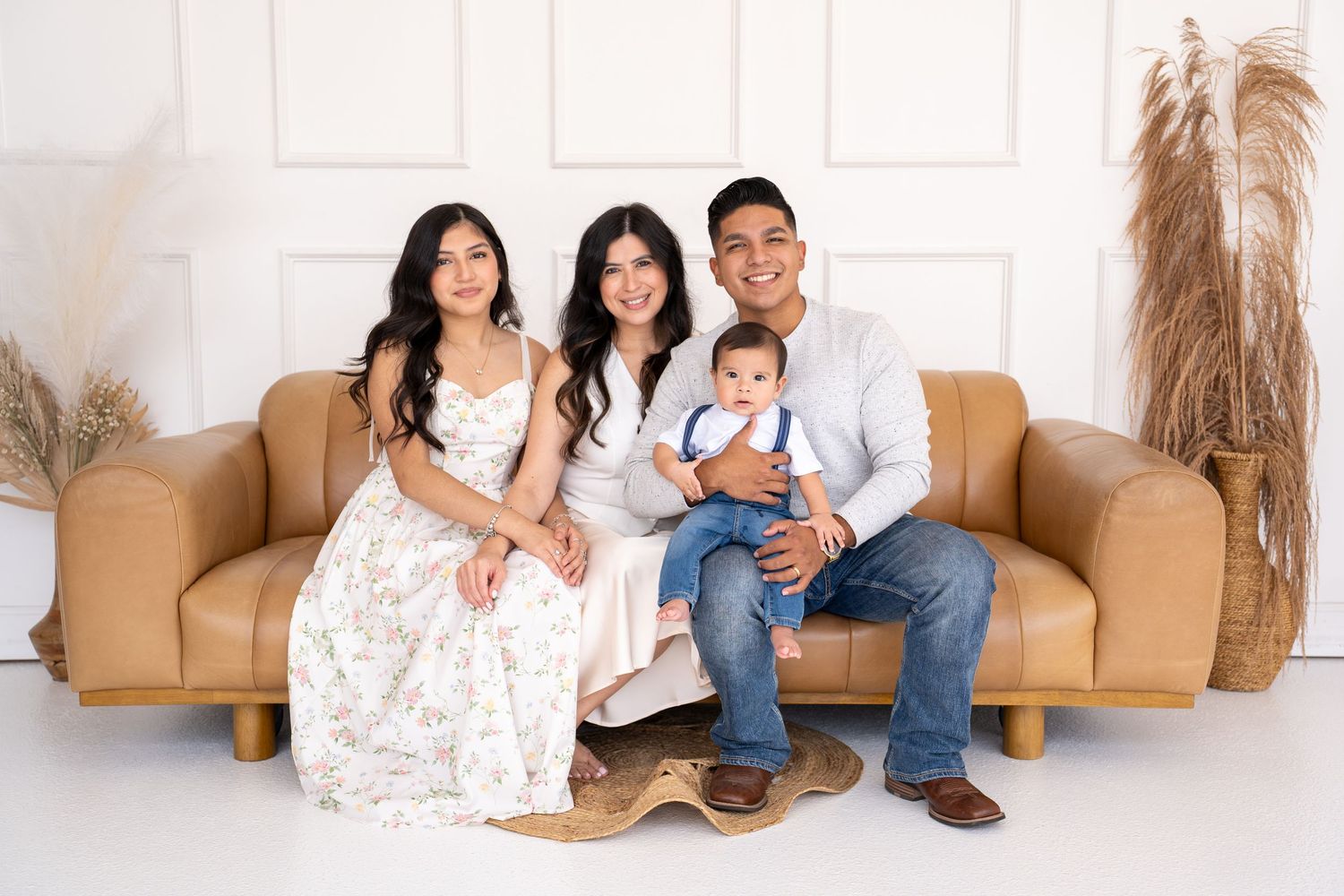 Family of four sitting on white floor, parents with young daughter and baby boy, smiling warmly in bright studio setting.