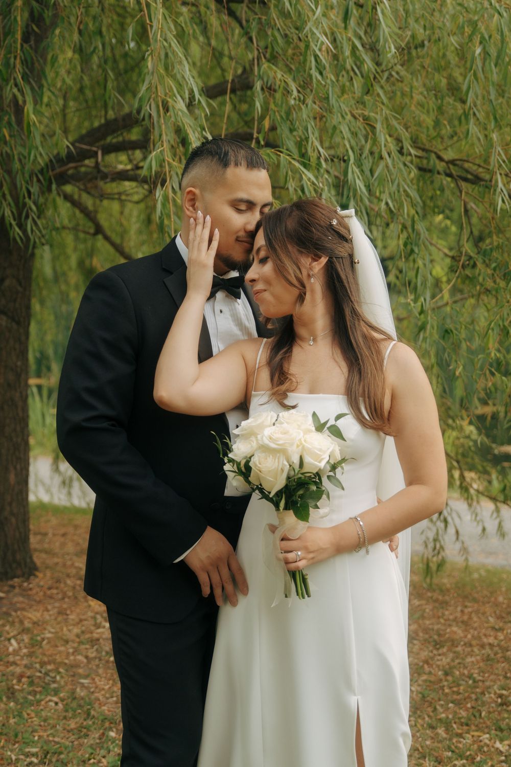 Bride and groom share a romantic moment under a willow tree, bride holding white rose bouquet in elegant white gown.