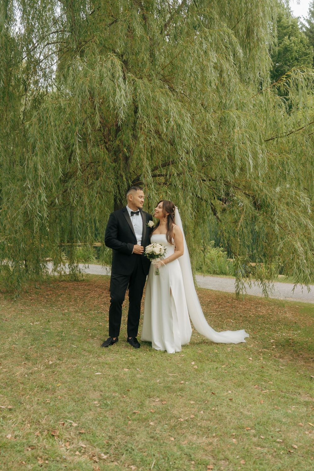 Bride and groom share a romantic moment under a weeping willow tree, bride holding white bouquet in elegant gown.