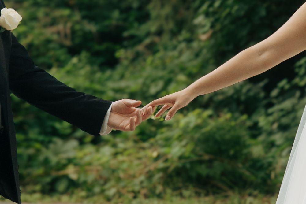 Bride and groom gently holding hands outdoors with lush green foliage in the background at a wedding.