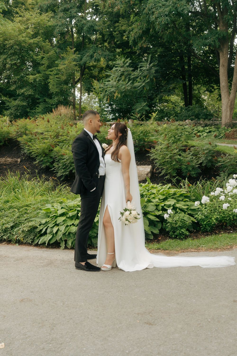 Elegant bride and groom share an intimate moment on garden path surrounded by lush green foliage and white flowers.