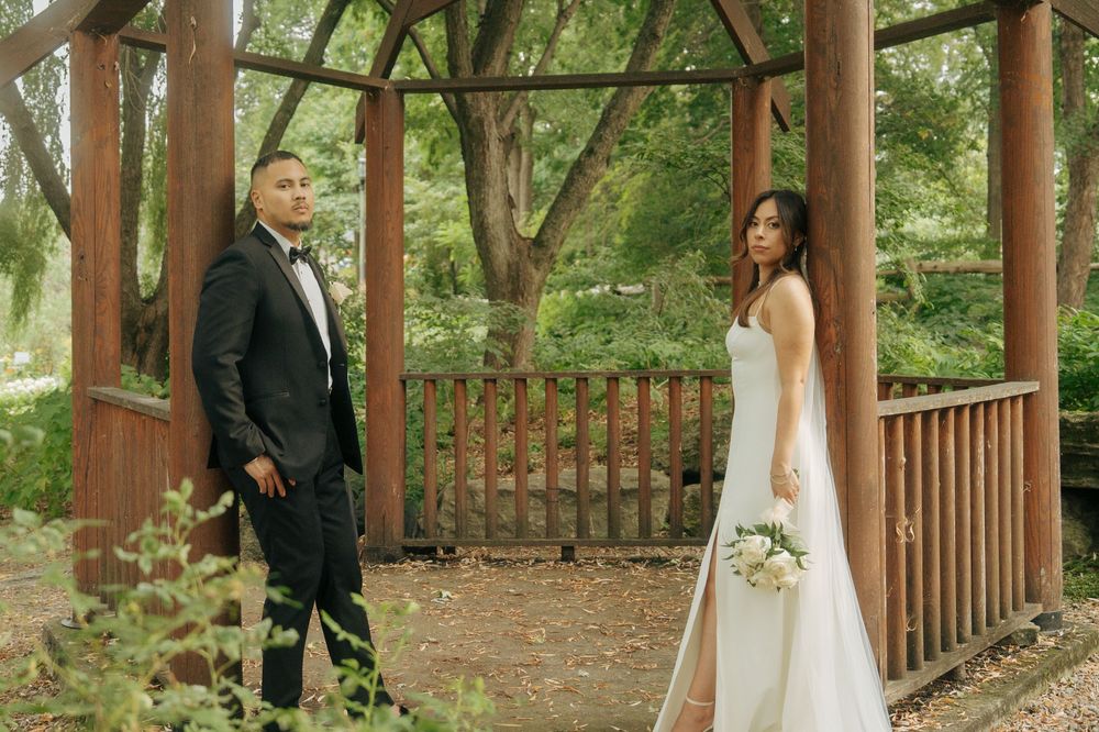 Bride in white gown holding bouquet and groom in black tuxedo pose under a wooden gazebo surrounded by lush greenery.