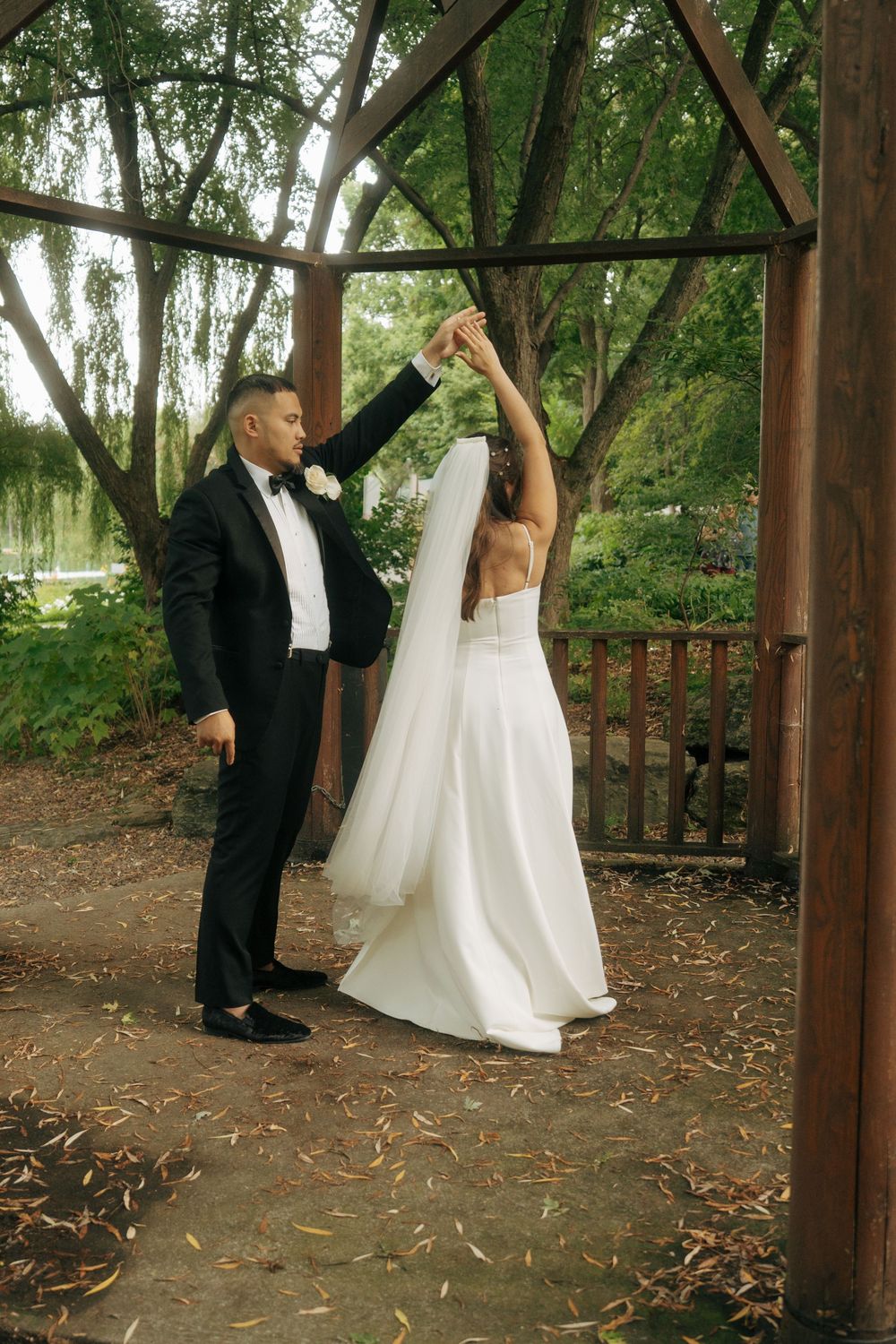 Groom in black tuxedo spinning bride in white wedding dress and veil under wooden gazebo in lush garden.