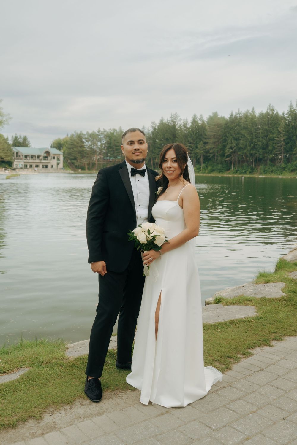 Bride and groom posing by a lakeside in elegant wedding attire, white gown and black tuxedo, holding a bouquet.