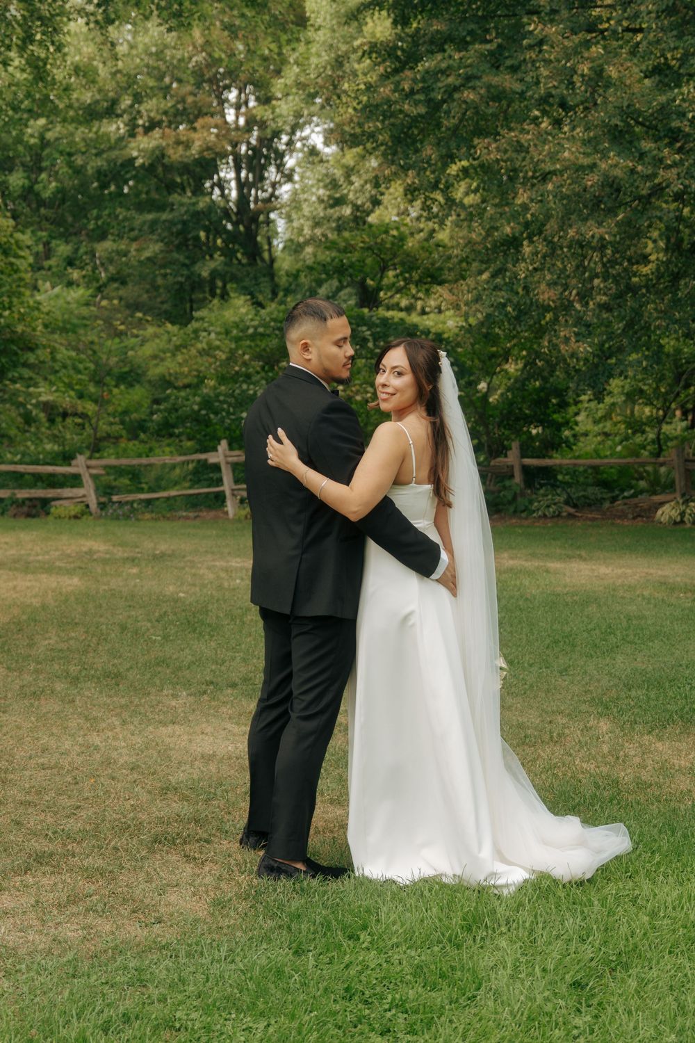Bride and groom embrace in a lush green garden, bride in white gown with veil looks back at camera.