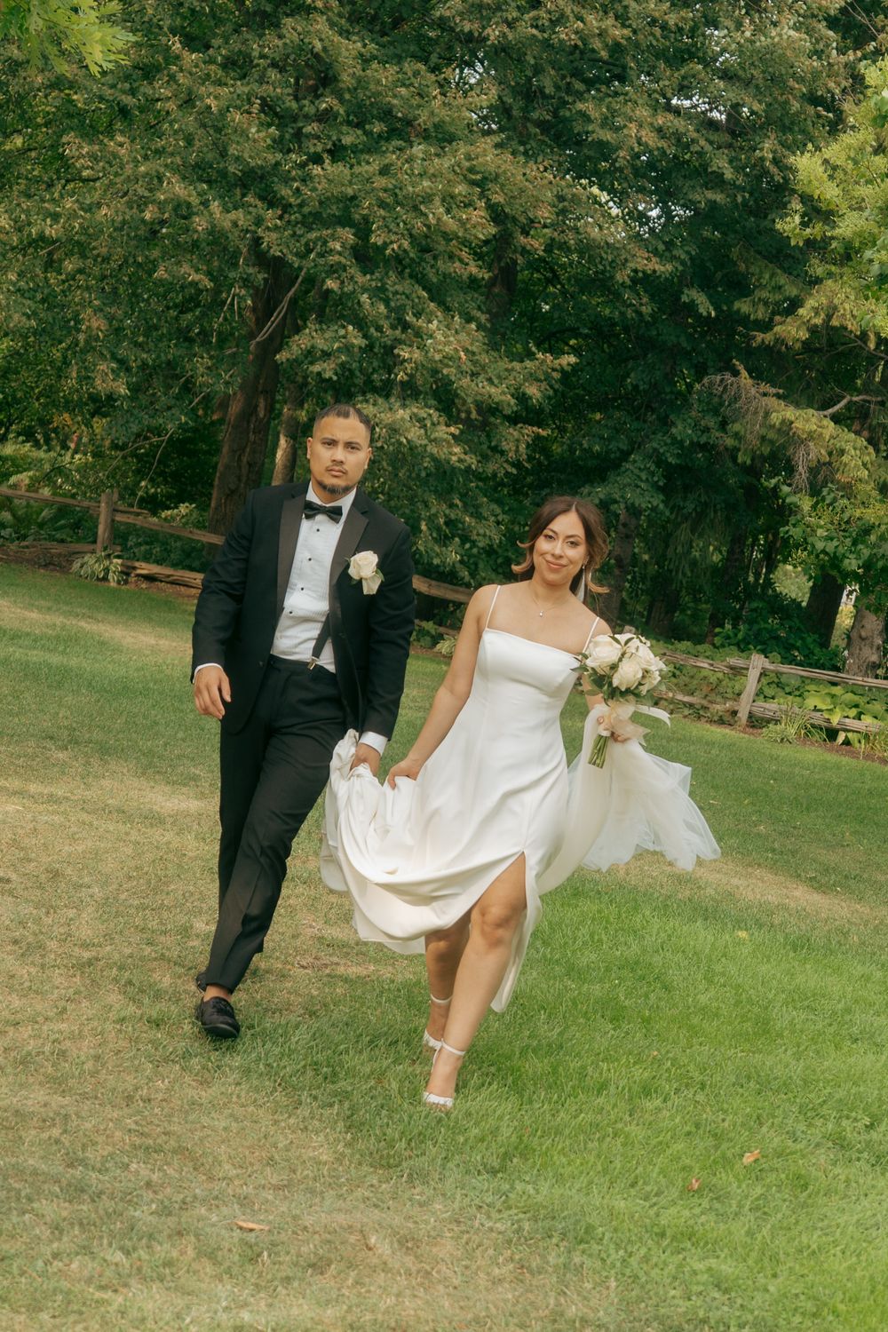Bride and groom walking hand in hand across green lawn, bride in white dress holding bouquet, groom in black tuxedo.