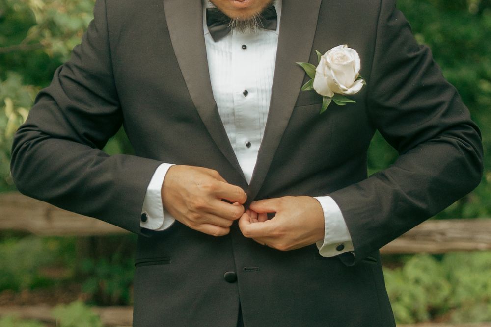 Groom in dark tuxedo with bow tie and white rose boutonniere buttoning jacket outdoors.