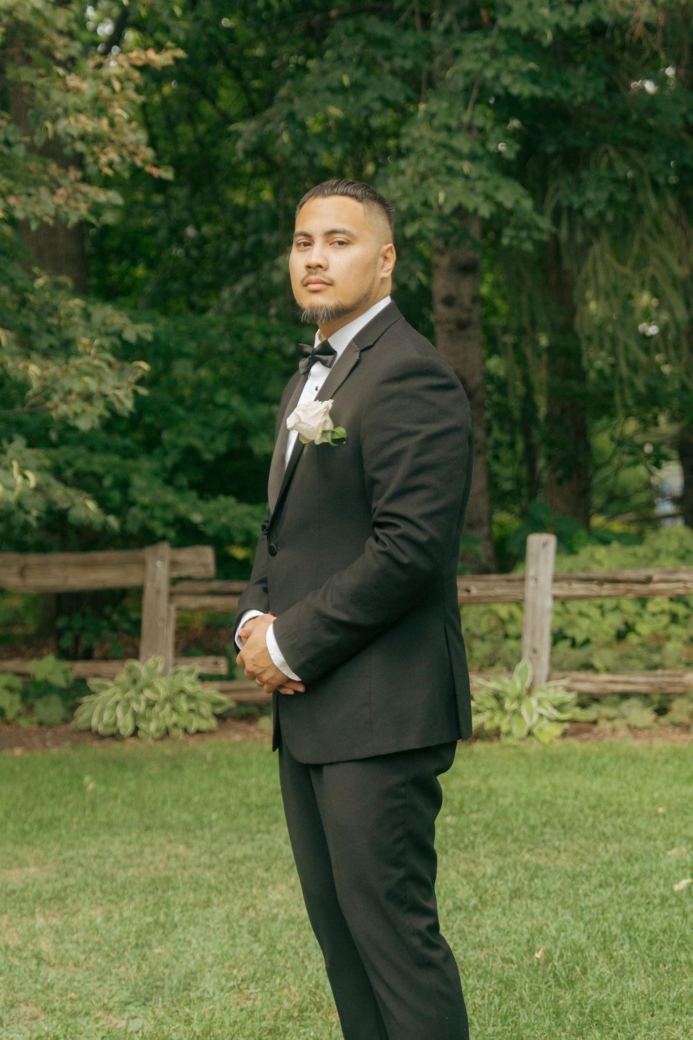A groom in a black tuxedo with boutonniere stands on green grass before a wooden fence and lush trees.