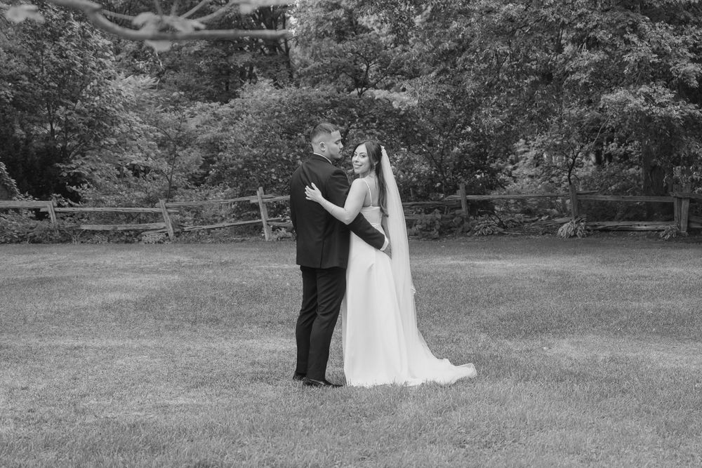 Black and white photo of a bride and groom embracing on a lush green lawn with trees and a fence backdrop.