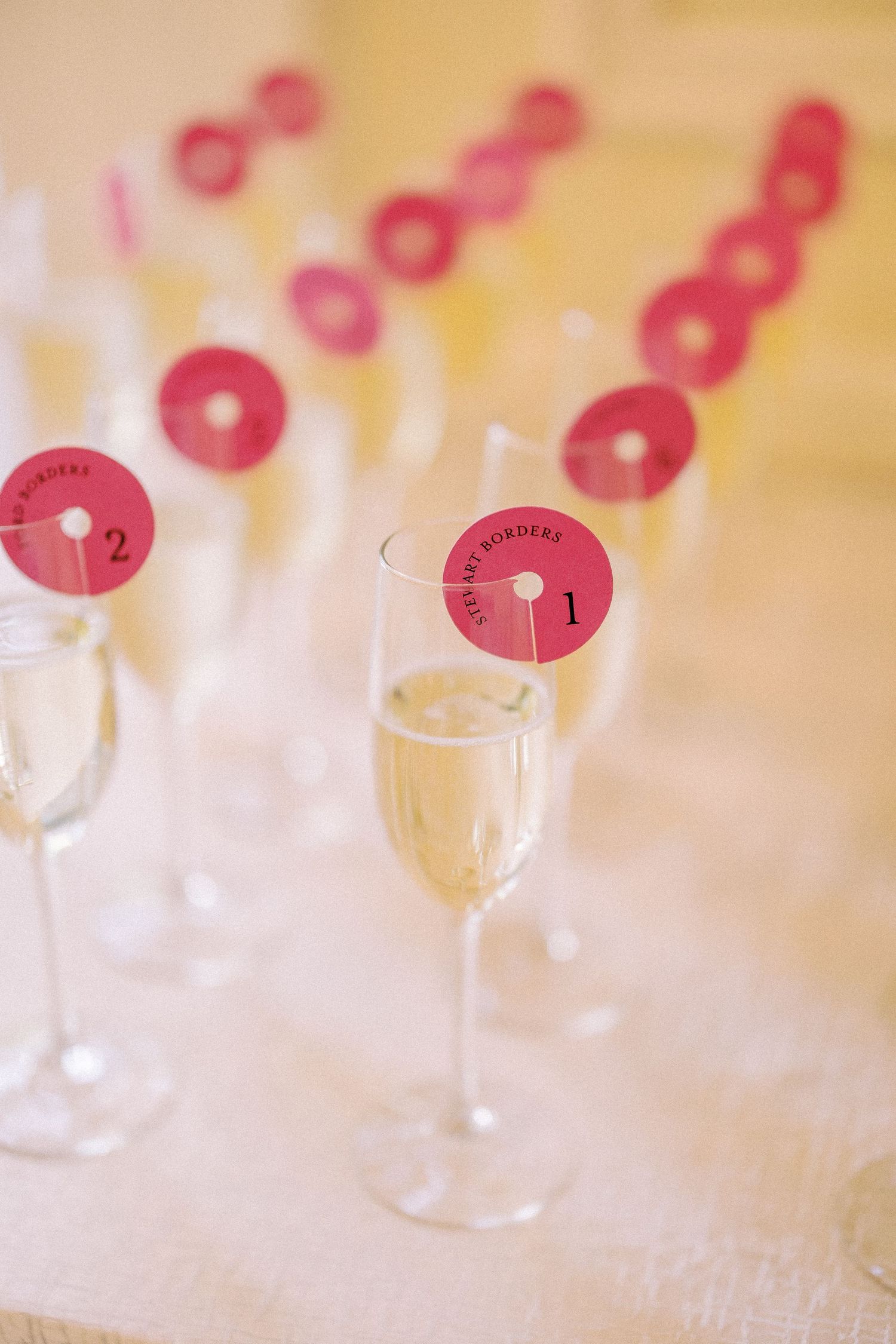 Champagne flutes with pink circular number tags on sticks for wedding seating assignments on a white table.
