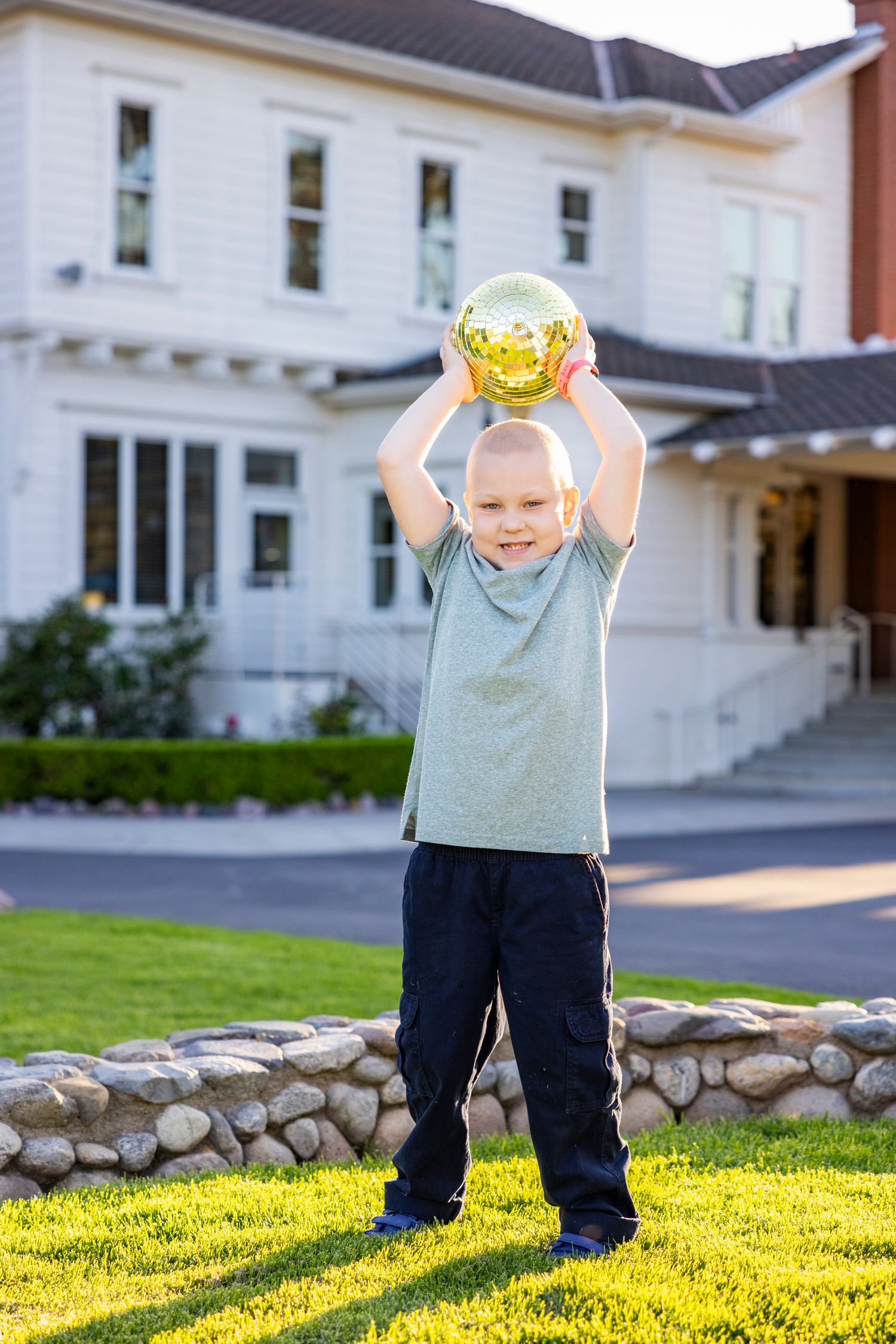 Young boy holding a shiny gold ball above his head on a green lawn in front of a white house.