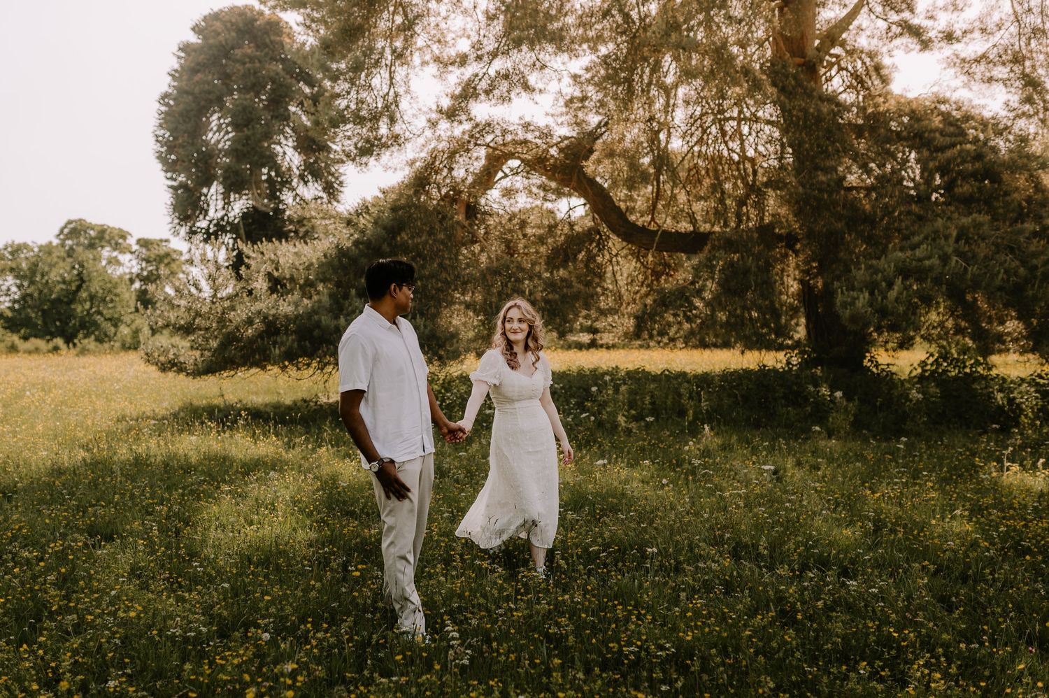 Couple holding hands walking through a sunlit meadow with lush green trees, man in white shirt and woman in white dress.