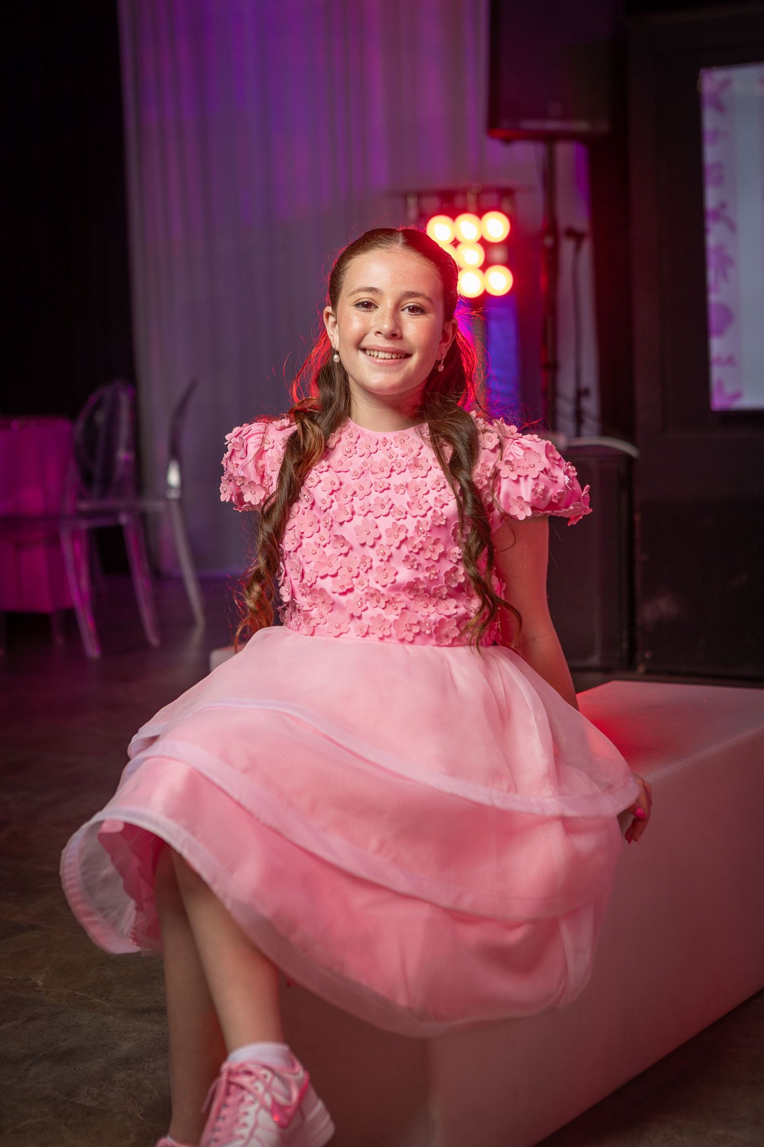 Young girl in pink floral dress smiling at a party event with colorful stage lights in background.
