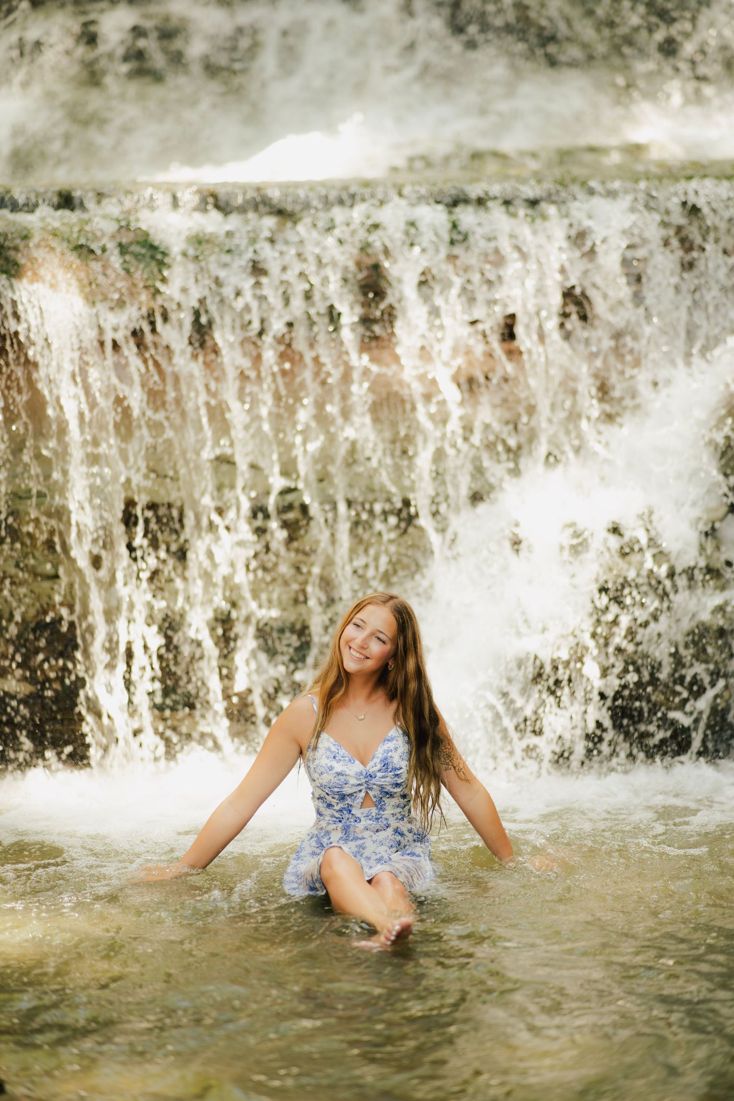 Young woman in blue floral dress sitting in shallow water before a cascading waterfall.
