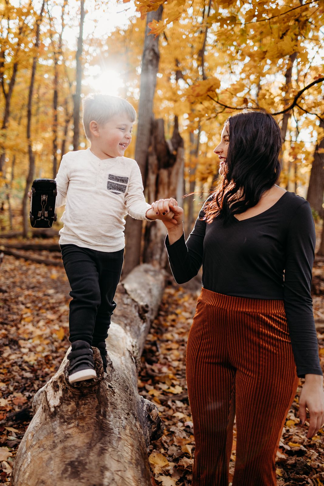 A mother and son walking through the forest. Her son walks across the log and they are smiling at each other. The sun is shining in the back