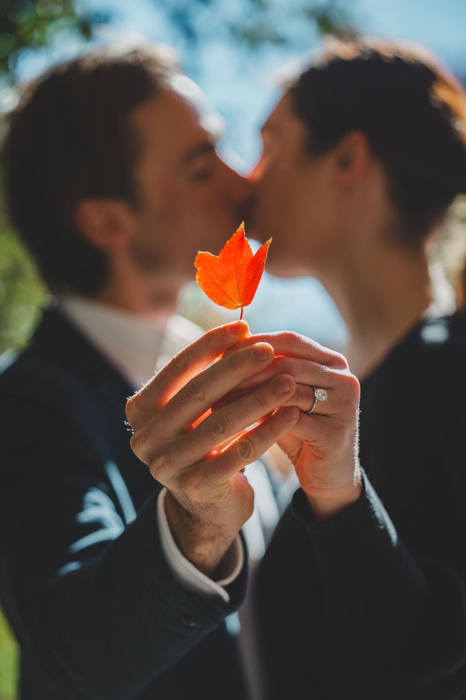 Couple kissing behind a vibrant red autumn leaf held up, woman wearing engagement ring, romantic fall wedding photo.