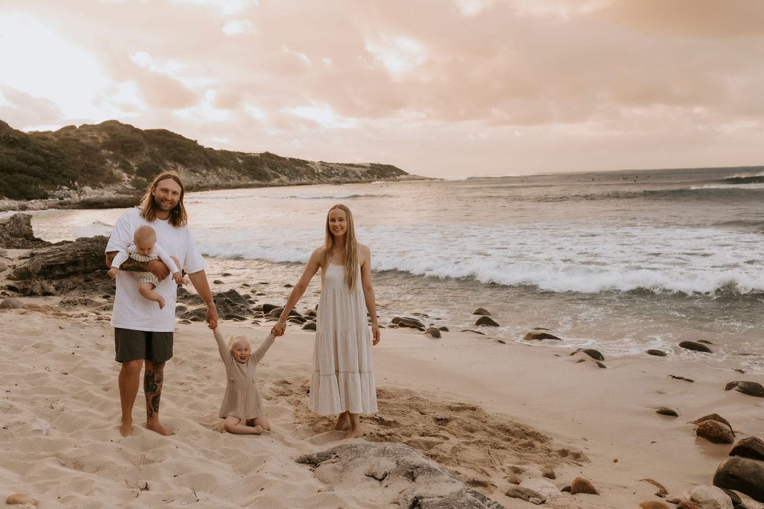 Family of four walking through a sunny wildflower field, parents holding toddler hands, golden hour light.