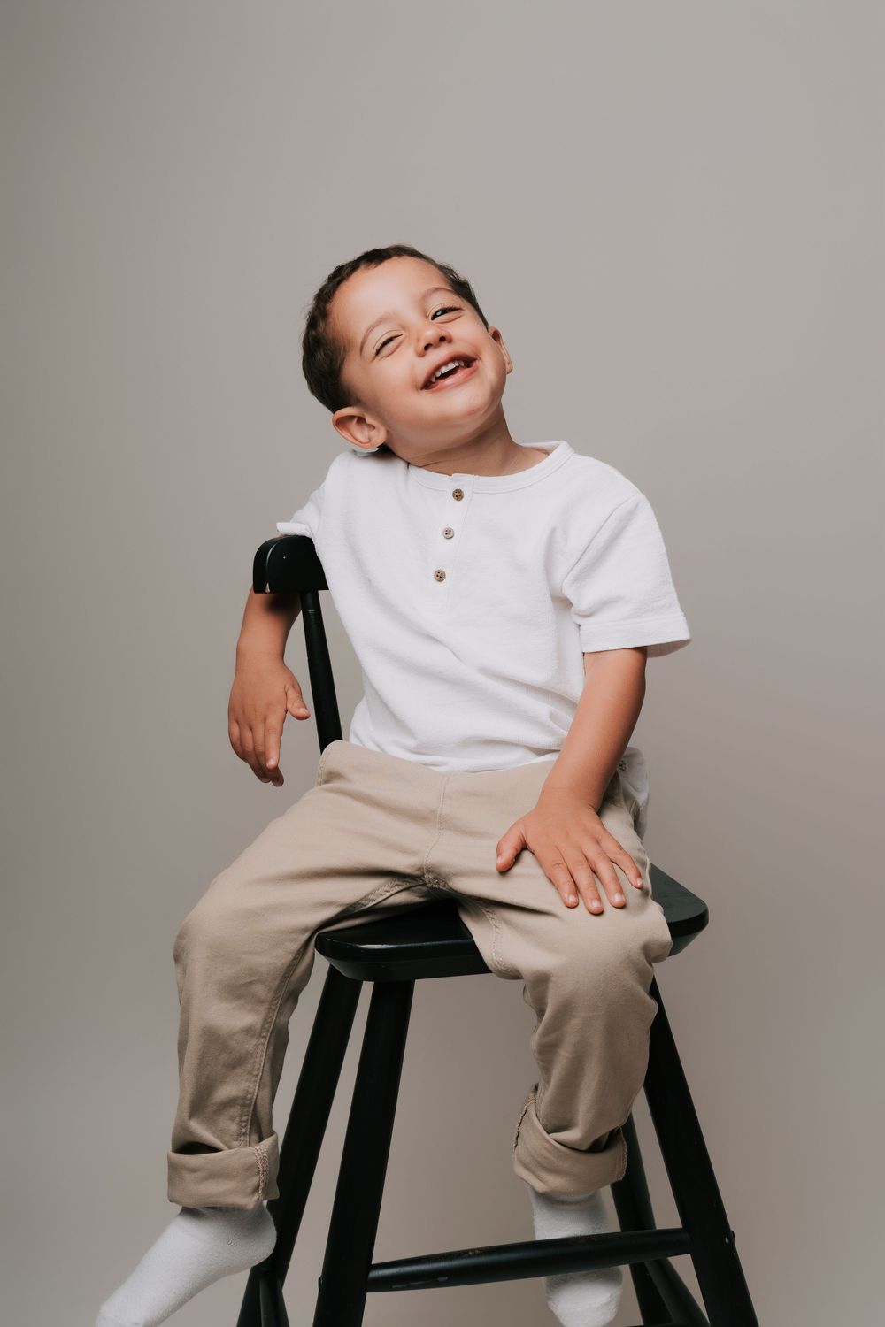 A cheerful young boy in a white henley shirt and khaki pants sits laughing on a black stool against a gray background.