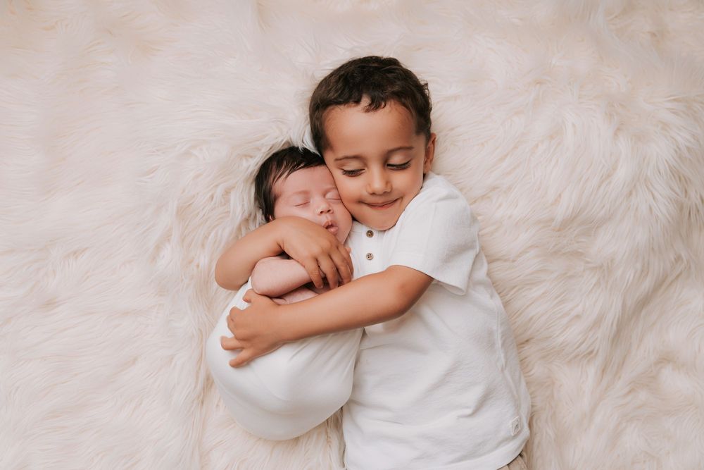 A young boy in white gently cradles a sleeping newborn on a soft cream fur blanket.