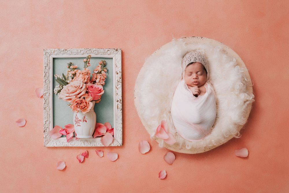 Newborn baby swaddled in white wrap sleeping in round basket beside floral arrangement on peach background.