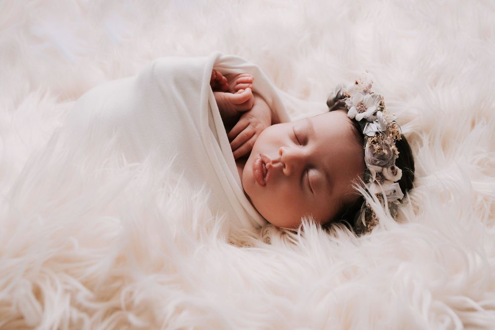 Peaceful newborn baby sleeping on fluffy white fur, wrapped in white fabric, wearing a delicate floral crown.