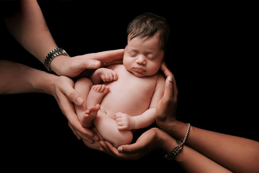 Newborn baby sleeping peacefully cradled in multiple hands against a black background.