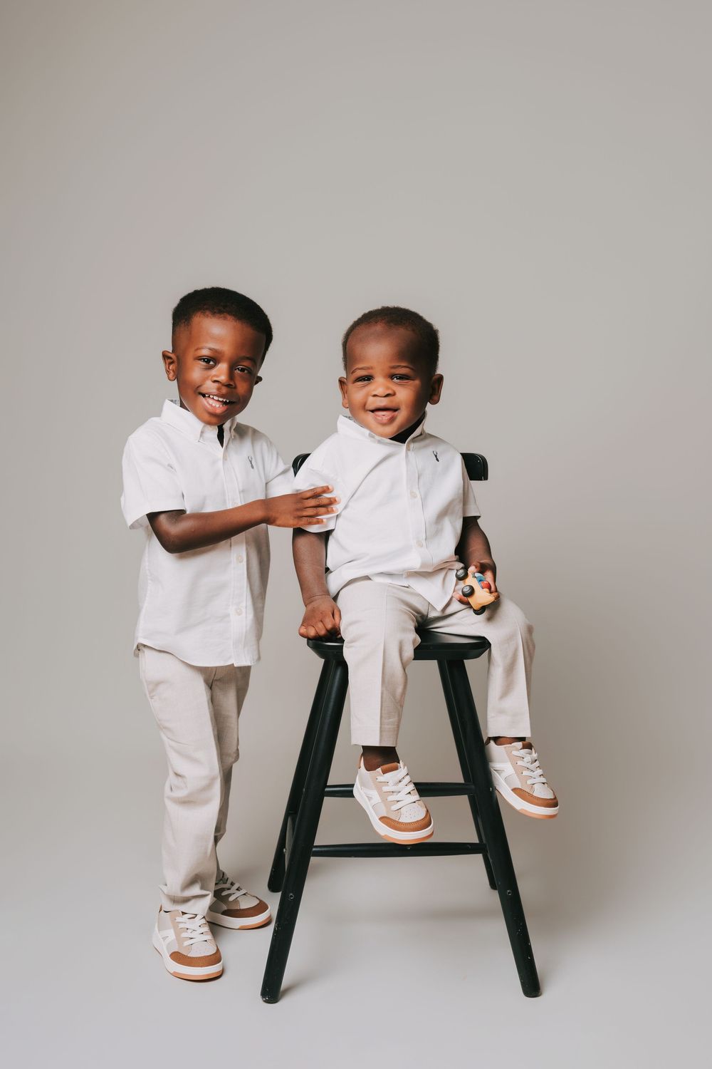 Two young boys in matching white shirts and khaki pants pose for a studio portrait, one sitting on a black stool.