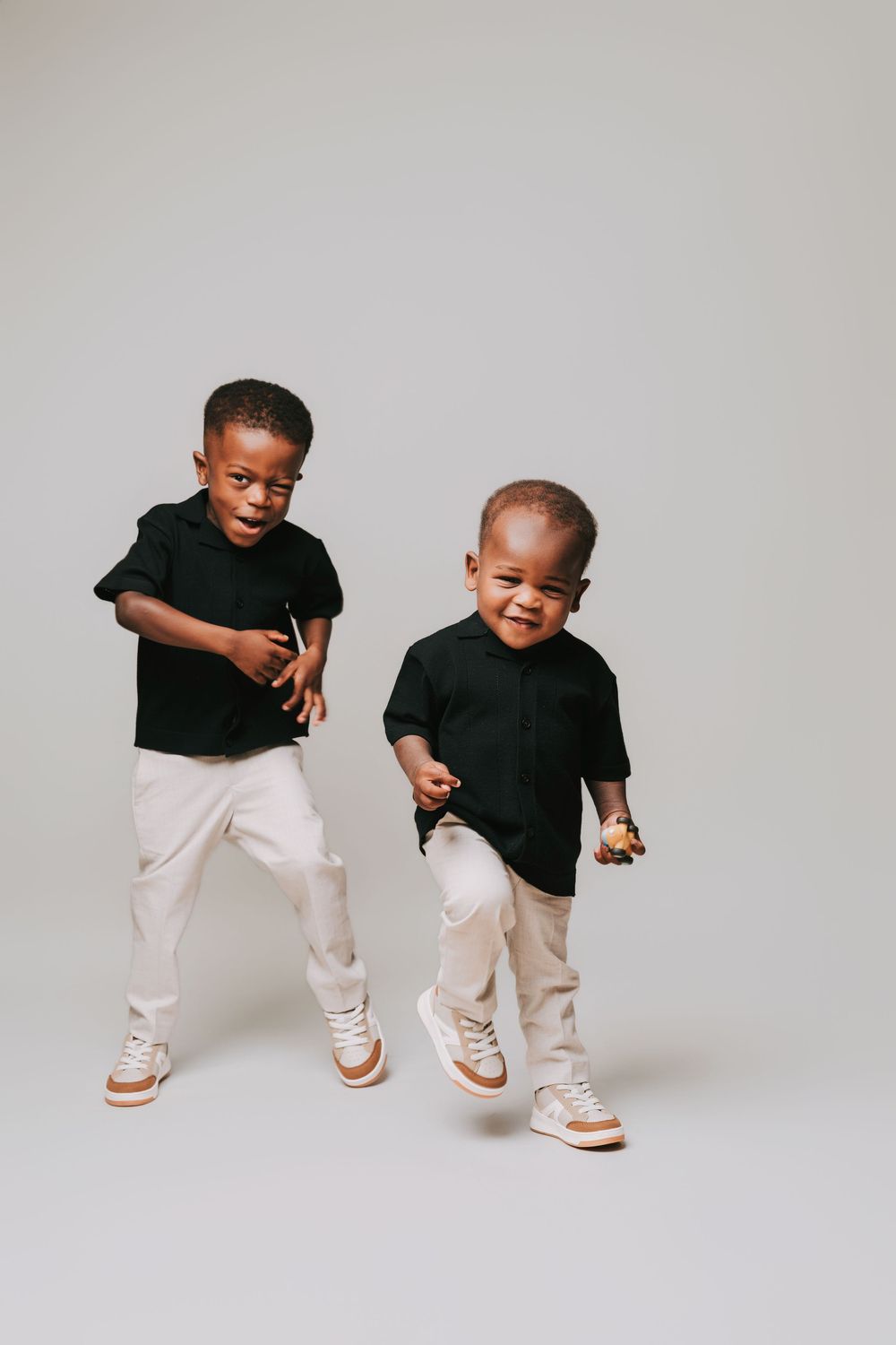 Two young boys in matching black shirts and beige pants pose playfully in a studio setting.