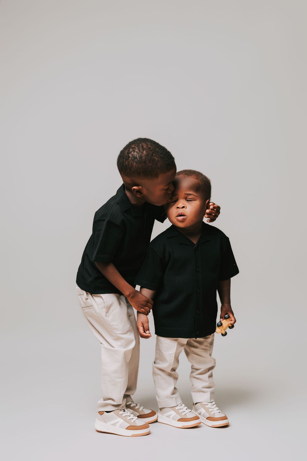 Two young boys in matching black shirts and beige pants, with the older child gently kissing the younger one on the cheek.