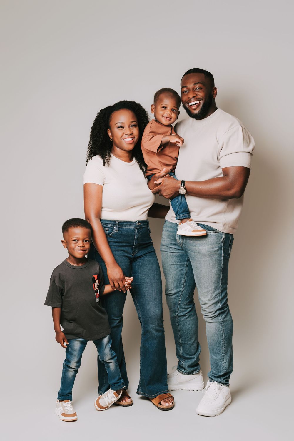 Happy Black family of four posing for a studio portrait, parents with two young children smiling against a gray background.
