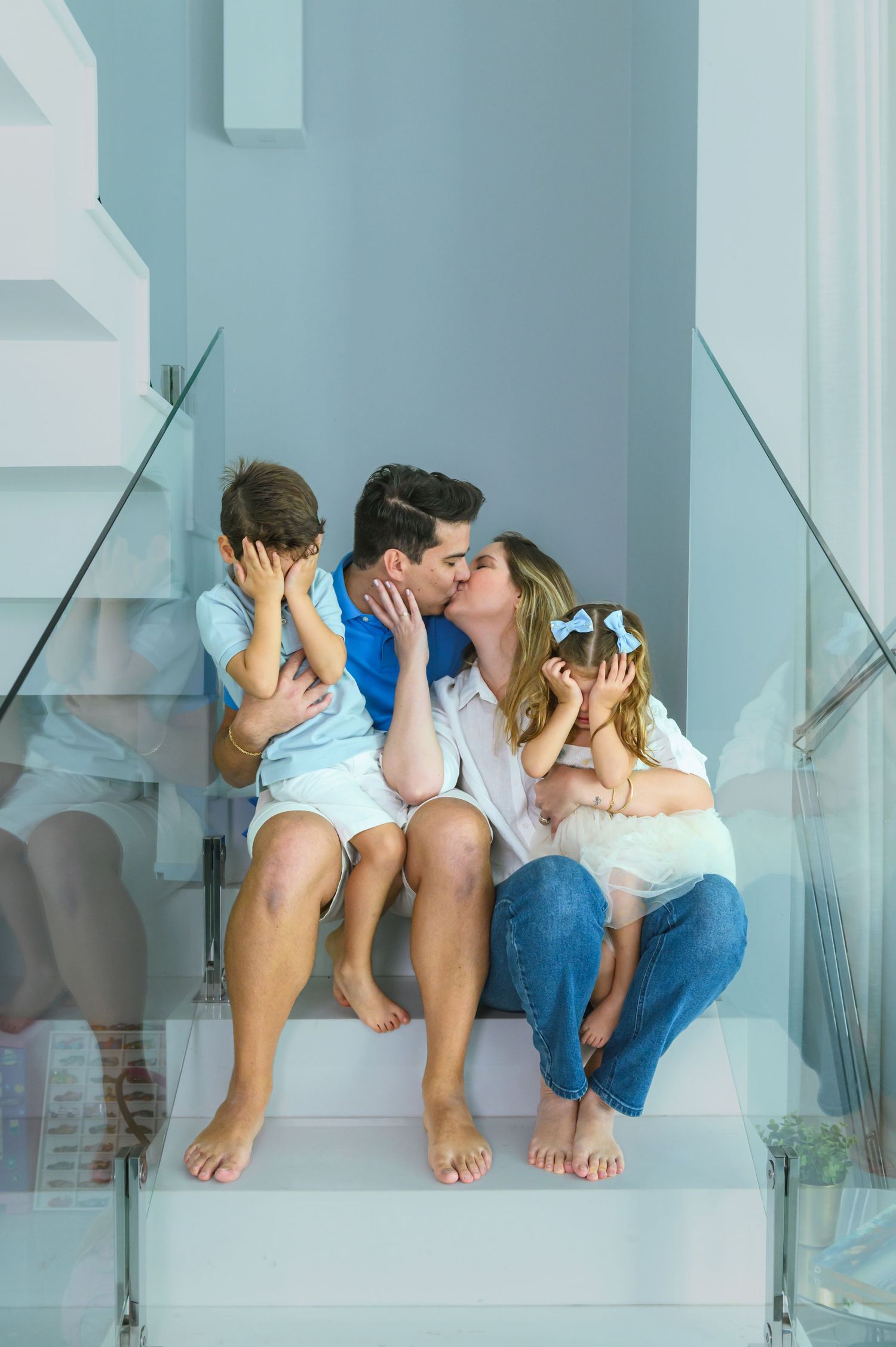 Parents kissing on stairs while two young children cover their eyes in a bright modern home.