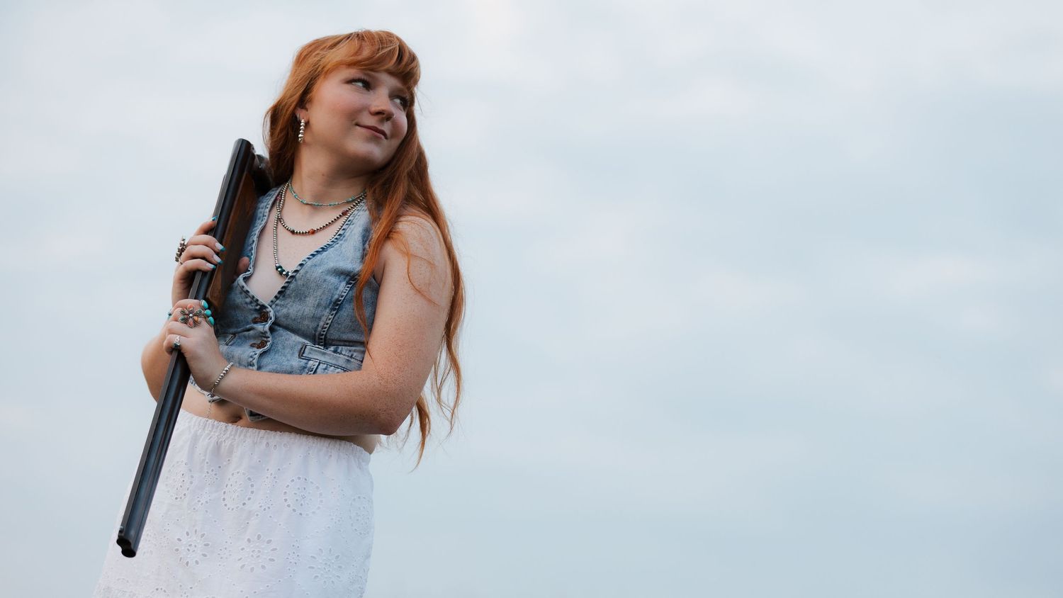 Young woman with long red hair in white dress holding a rifle, posing confidently against a cloudy sky.