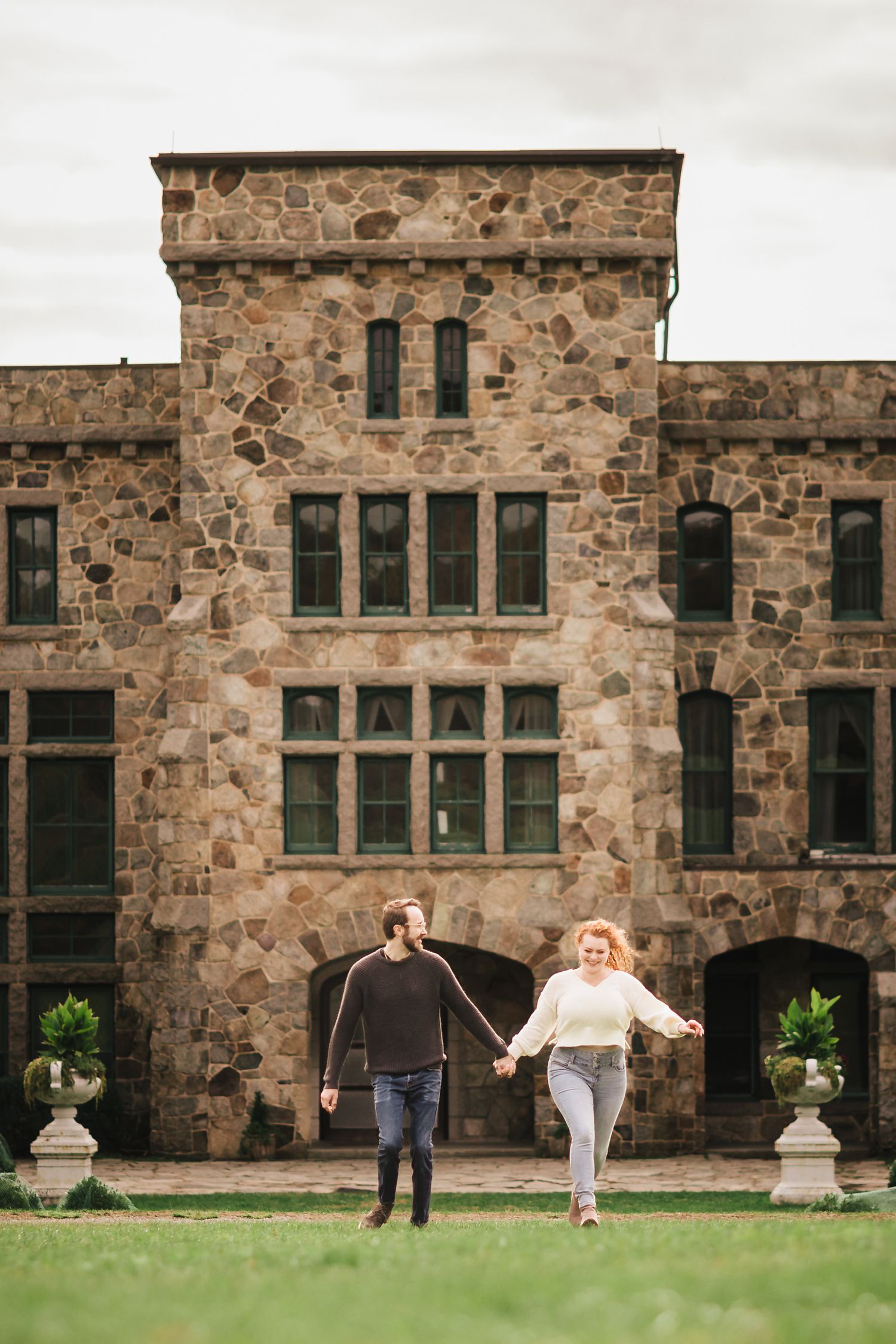 Engaged couple running hand in hand across the lawn in front of the historic stone Ames Mansion at Borderland State Park, laughing together.