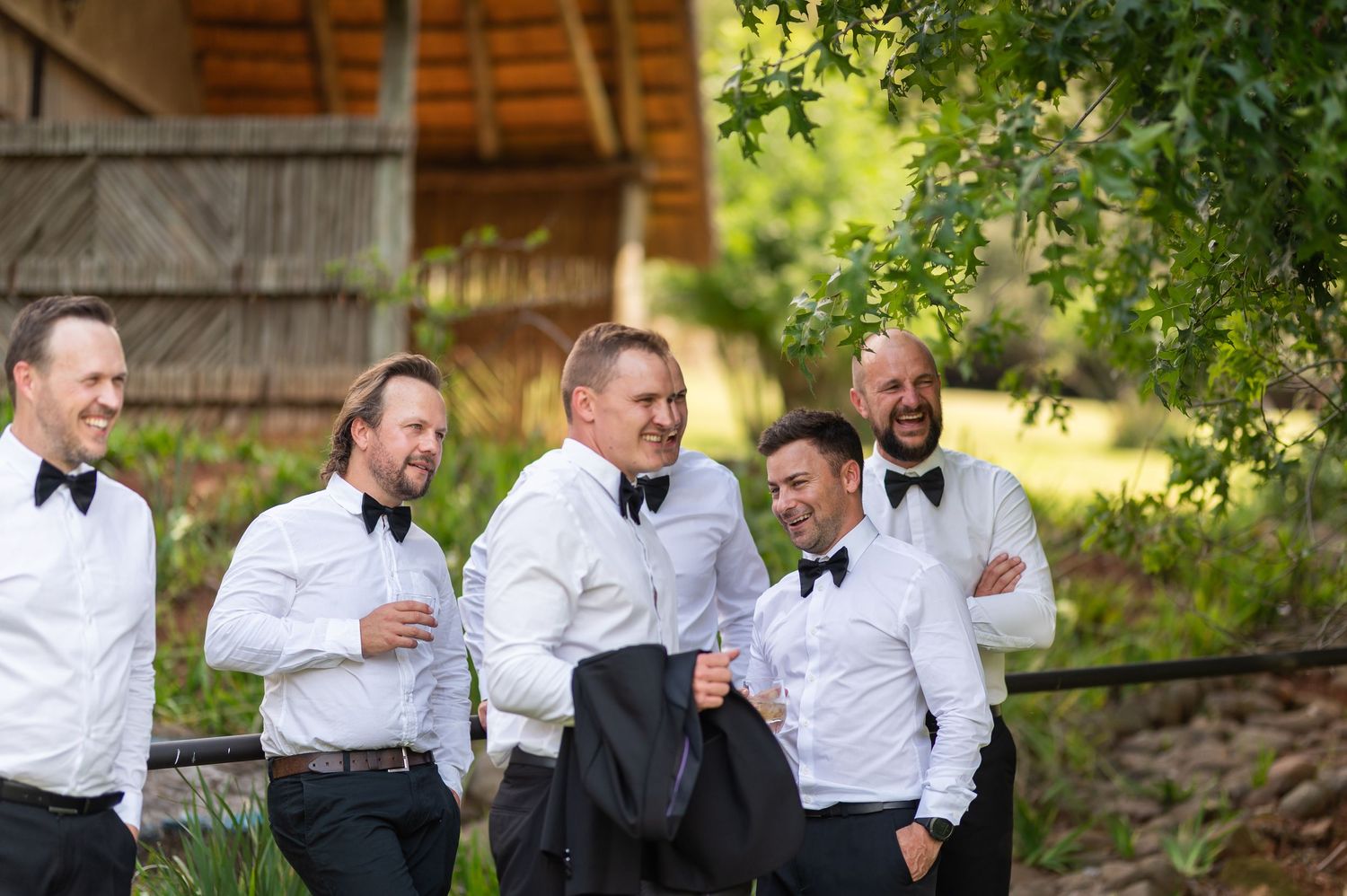 Five groomsmen in black tuxedos and bow ties laughing together outdoors near a rustic wooden building.