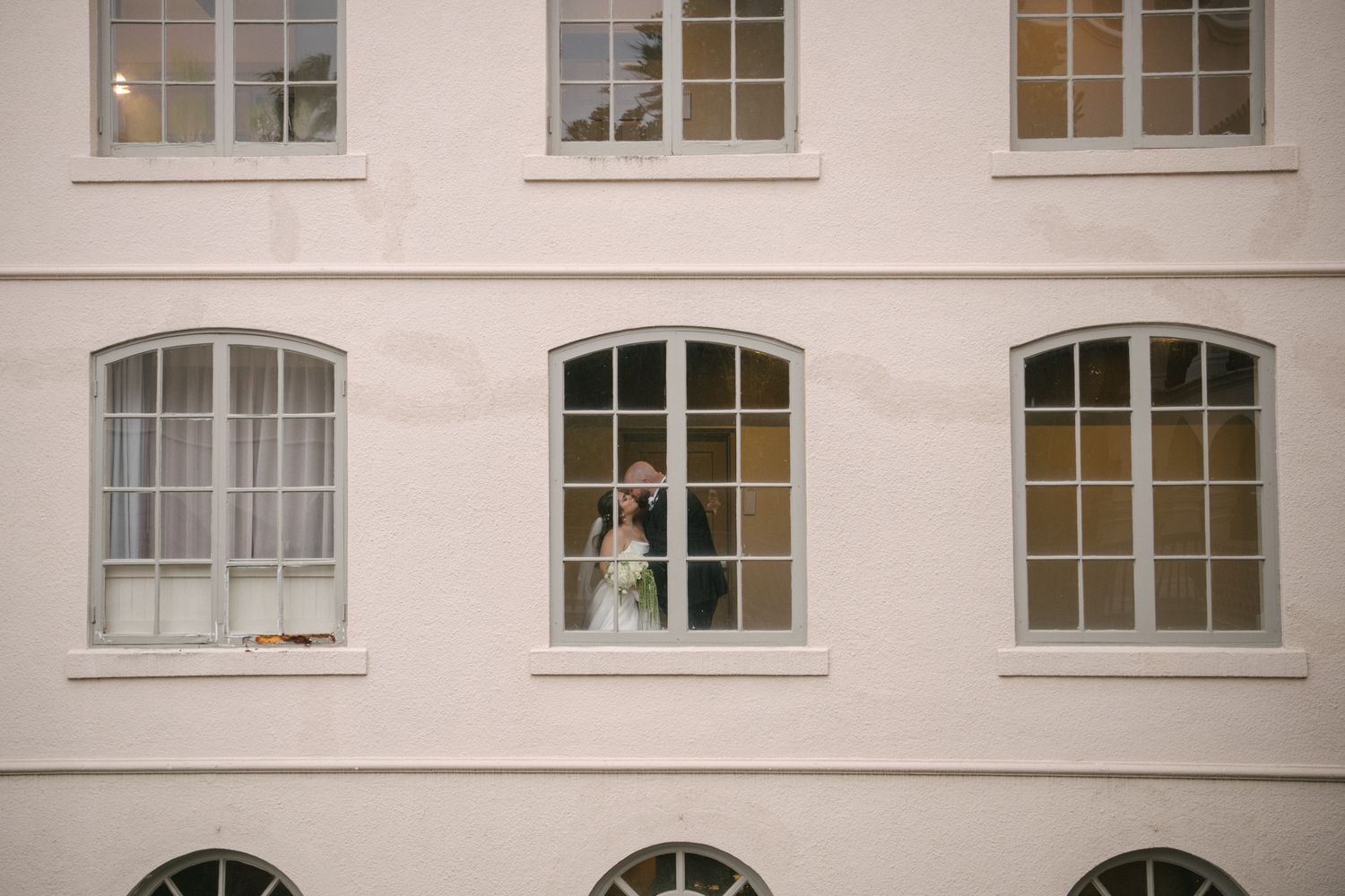 A couple in wedding attire embraces in front of an elegant white stone building entrance with blue-green doors.